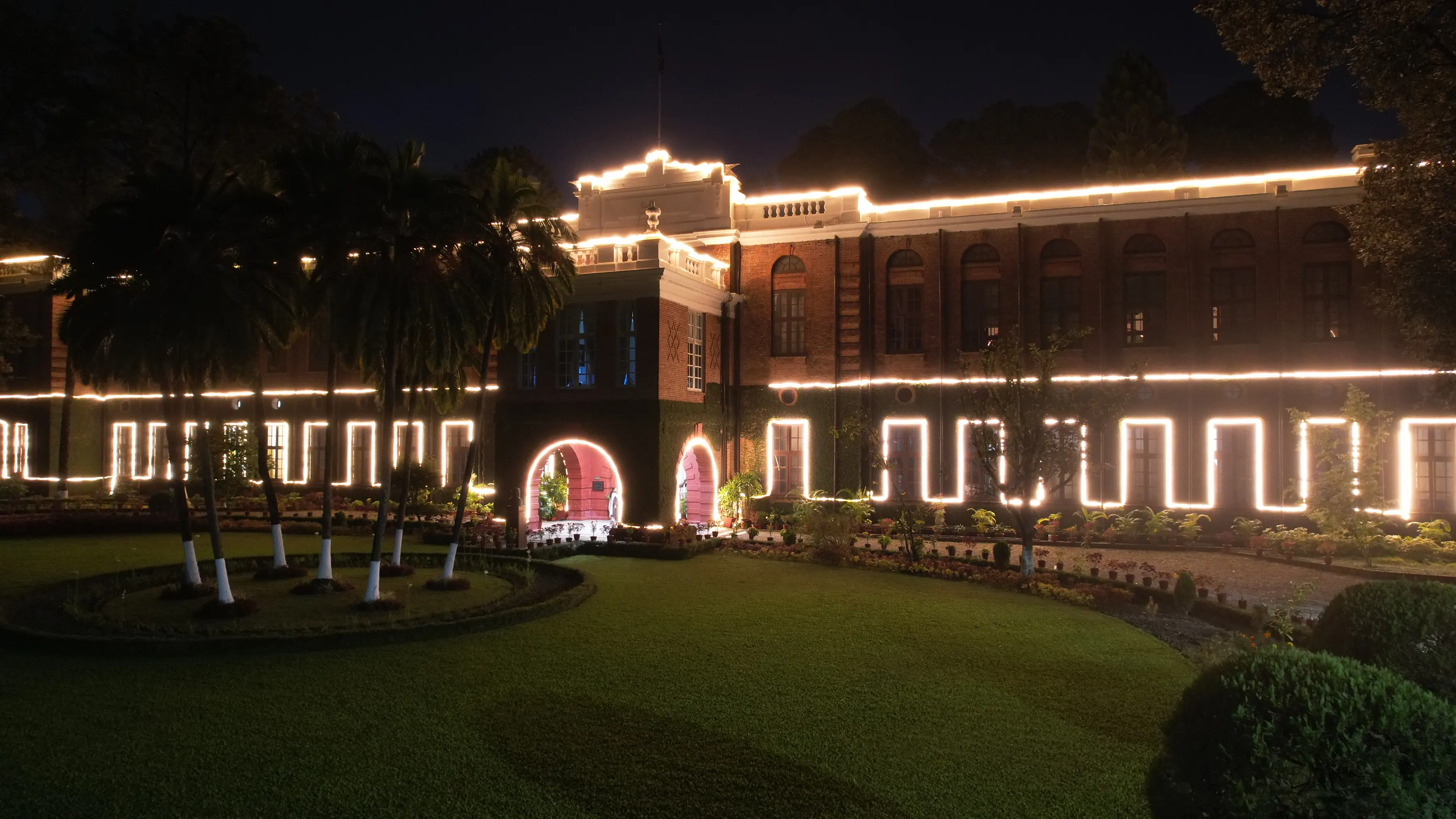 The Main Building at night, its arches, roofline and entrance outlined in lights, seen across the circular garden