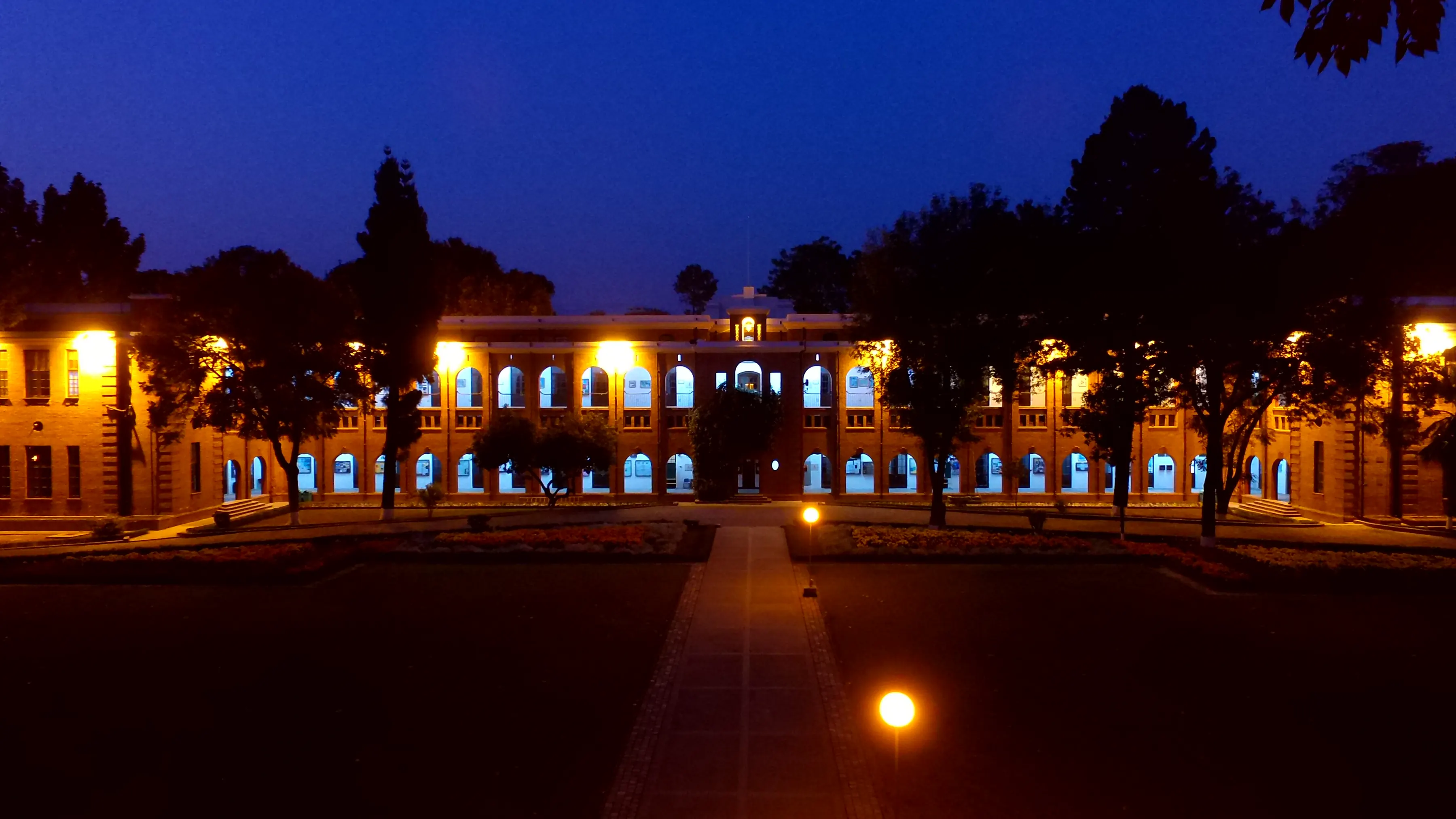 The Main Building at night, its windows and arches lit, seen down a lamplit path across the Gurus Lawn