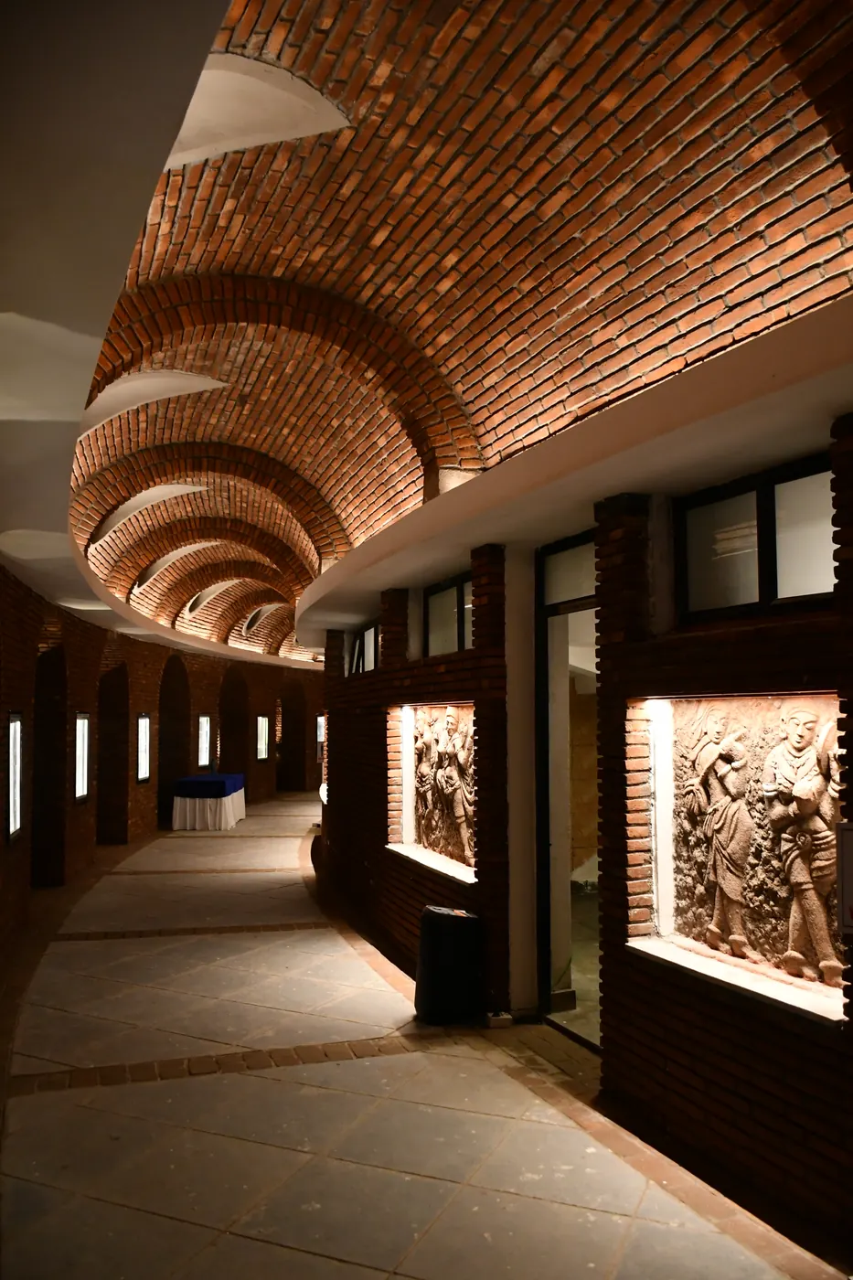 A curving corridor under a vaulted brick ceiling, lit relief panels set into the brick wall